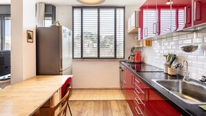 A compact kitchen with a wooden table, a refrigerator, red cabinets, a window with blinds, and a stainless steel sink. - Allenby Artsy Stay by Sea N' Rent (Tel Aviv)
