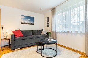 Moderni olohuone, jossa on harmaa sohva, pieni pyöreä pöytä, valkoinen matto ja suuri ikkuna, jossa on läpikuultavat verhot.
