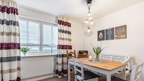 Un coin repas avec une table en bois, des chaises blanches, un radiateur et une fenêtre avec des rideaux.