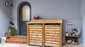 Une porte en bois, une porte noire ornée d’une fenêtre à motifs losangés, et une petite étagère avec des plantes et une lanterne.