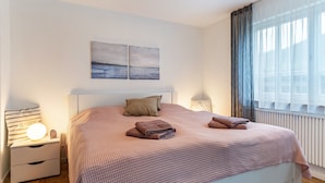 Un lit bien fait, avec une couverture à carreaux, un oreiller et des serviettes pliées. Au-dessus du lit, on peut voir deux tableaux accrochés au mur, une lampe de chevet et une table de nuit avec une lampe et un livre.