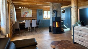 Interior rústico de una cabaña con chimenea de piedra, vigas de madera en el techo, comedor con sillas y cocina con armarios azules.