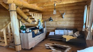 Interior de una cabaña de troncos con una escalera de madera, un sofá, un comedor con encimera negra y una lámpara colgante.
