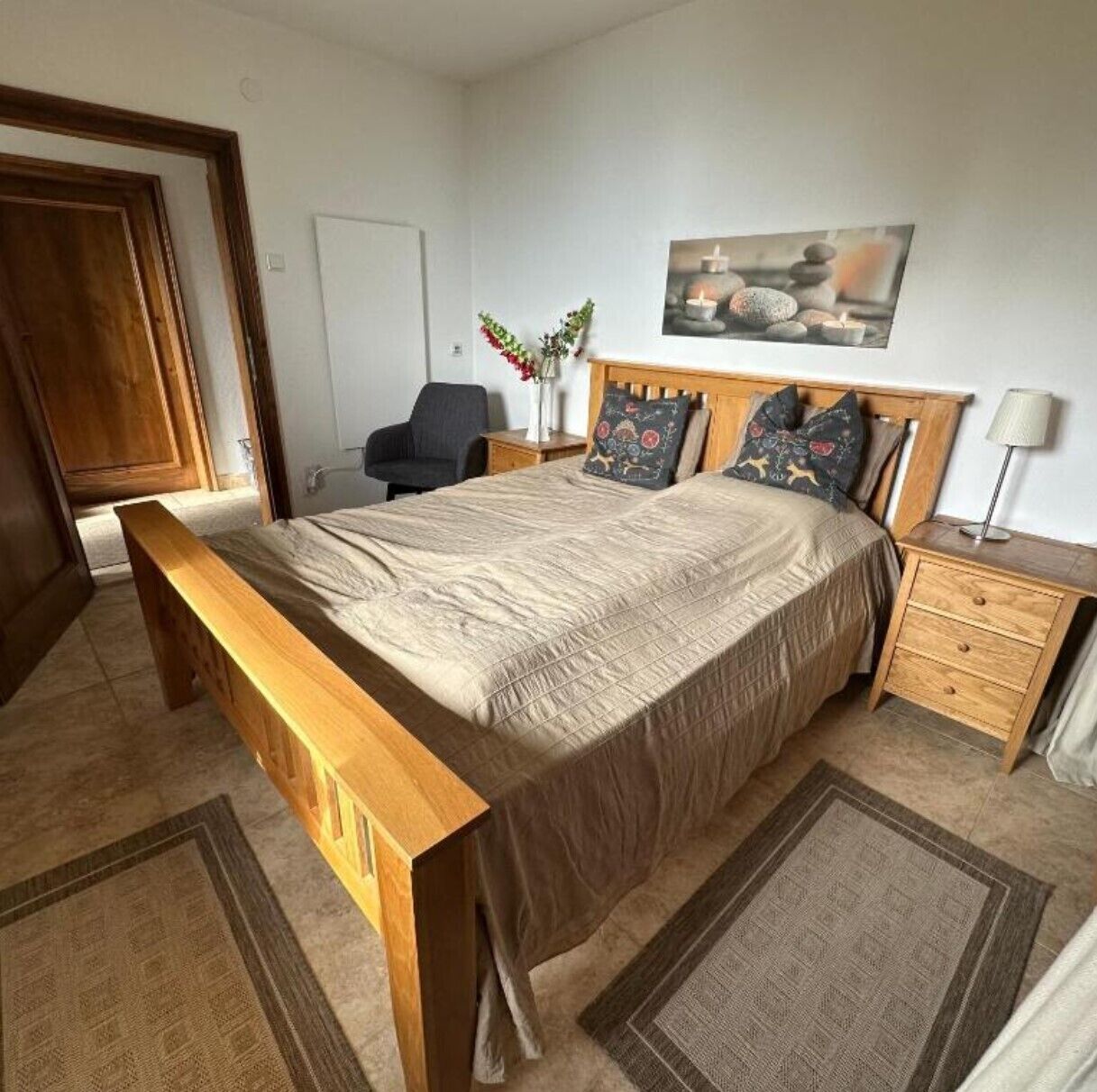 A bedroom with a wooden bed, a nightstand, a chair, a painting, and a door.