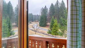 Vista desde una ventana con balcón de madera, que da a una zona boscosa con carretera y puente.