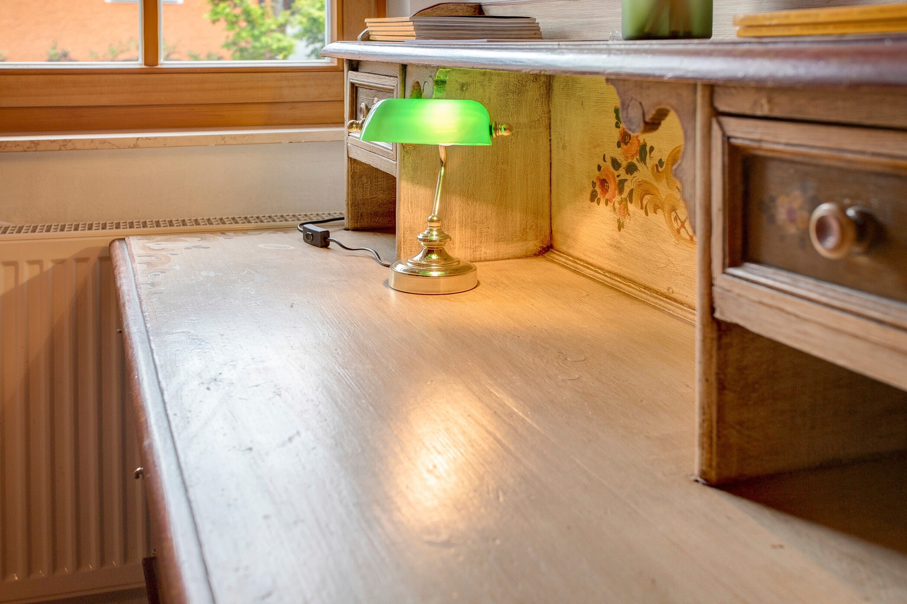Uma luminária de mesa com cúpula verde sobre uma escrivaninha de madeira, perto de um radiador.