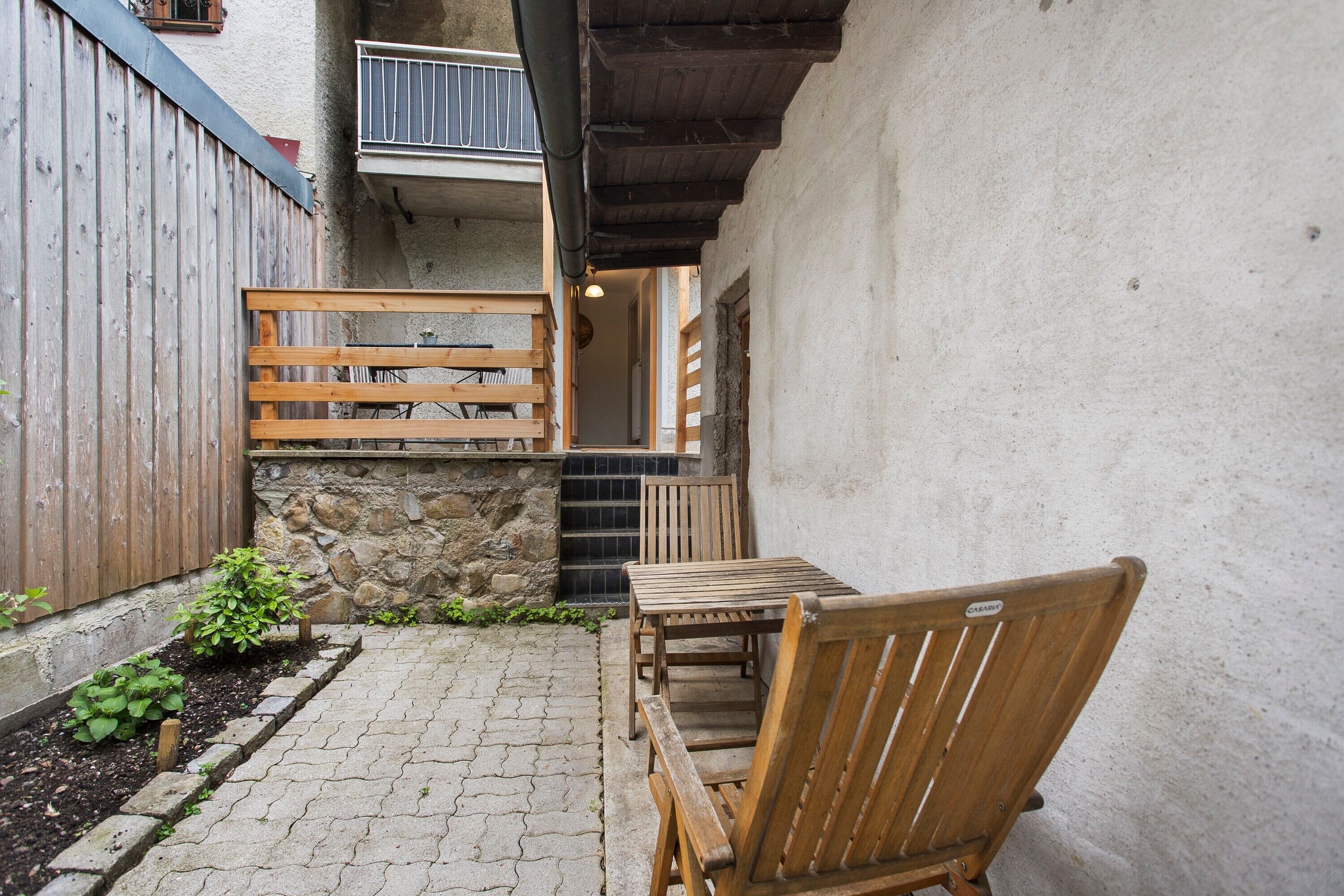 Un banc et une table en bois se trouvent dans une cour bordée d’un mur de pierre et d’une clôture en bois.