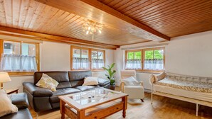 A cozy living room with a wooden ceiling, a leather sofa, a glass coffee table, a white armchair, and a daybed.