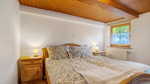 A bedroom with a wooden ceiling, a bed with patterned bedding, a nightstand with a lamp, and a window with a view of greenery.