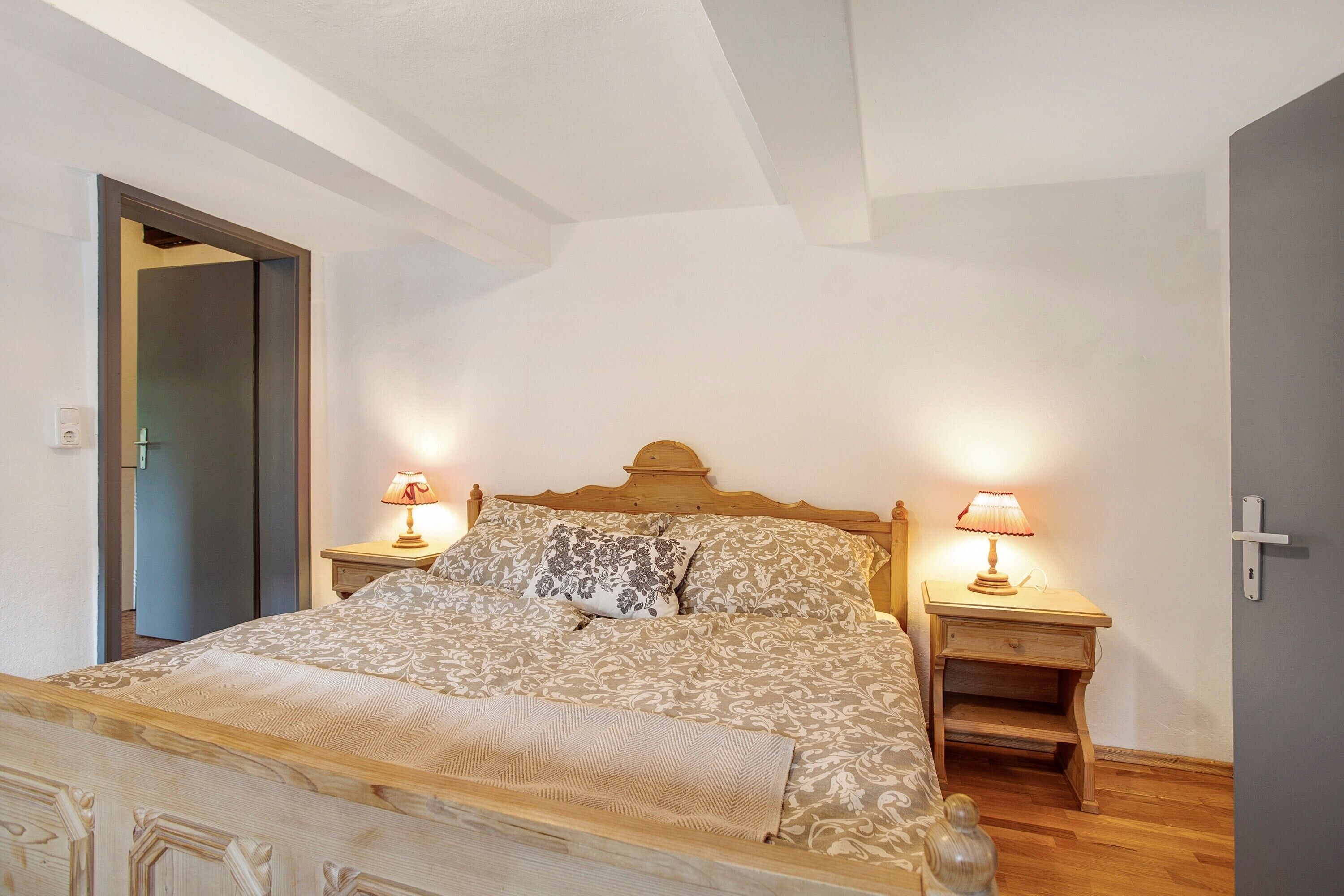 A bedroom with a wooden bed, two bedside tables with lamps, a door, and a wall-mounted light switch.