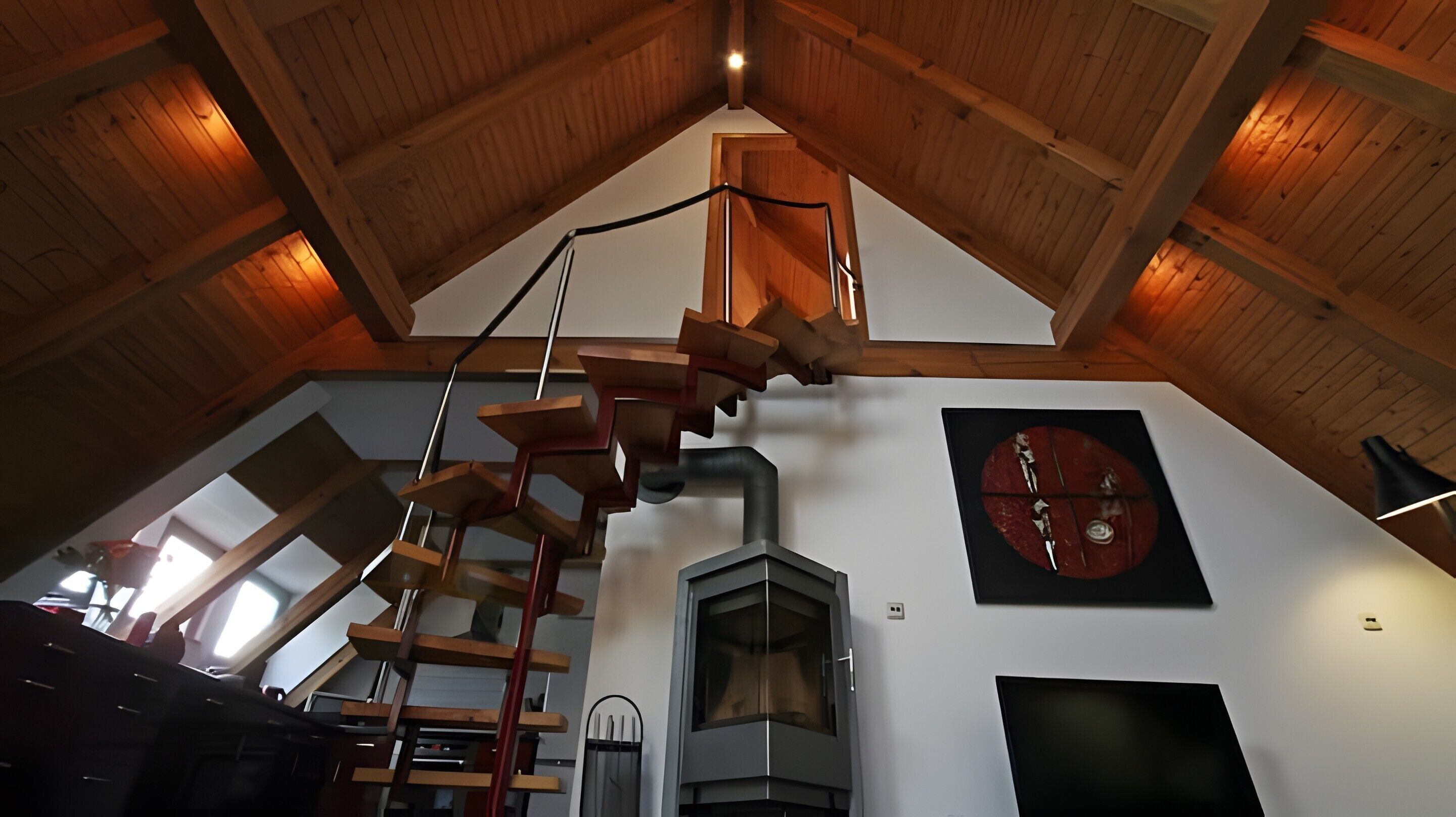 Une pièce aux hauts plafonds, avec un escalier en bois, une cheminée et une grande fenêtre.