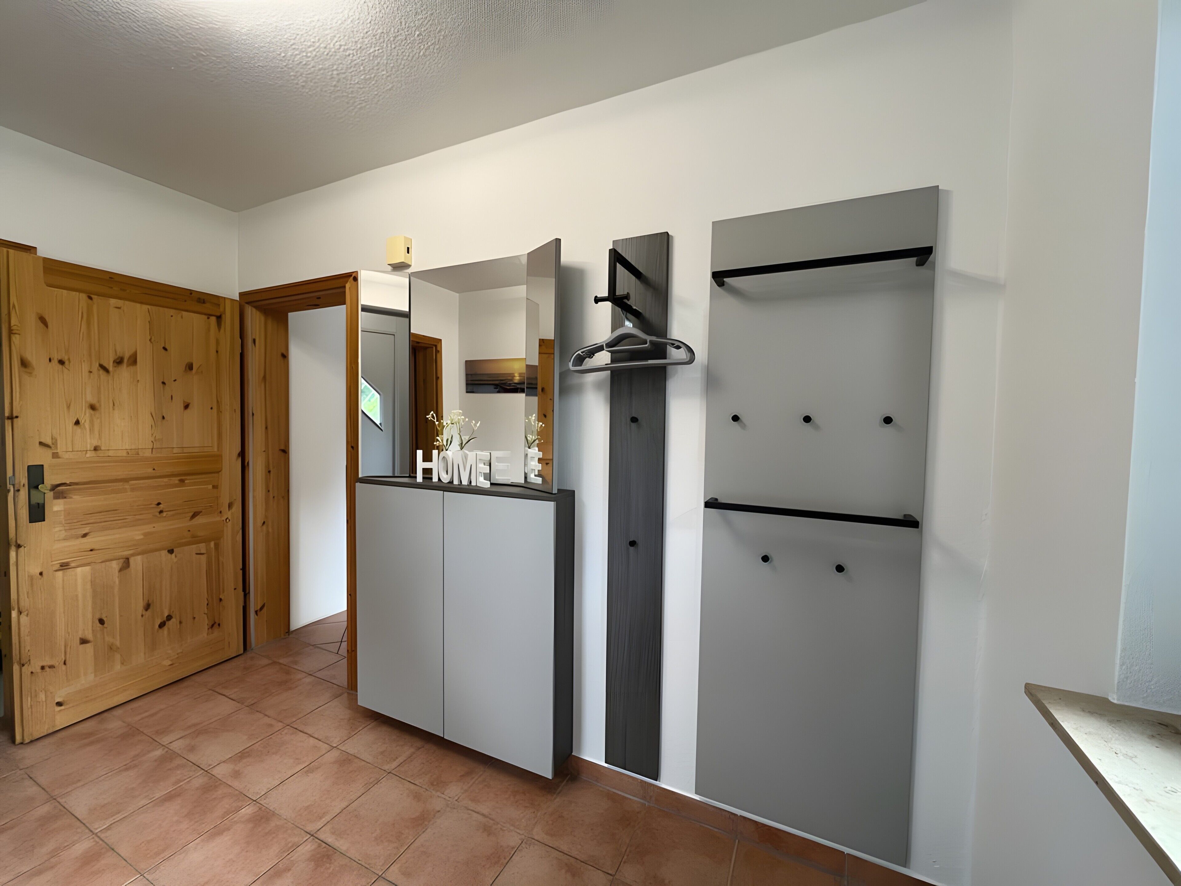 Una stanza con una porta in legno, un mobile grigio con uno specchio e una parete bianca con uno scaffale nero.