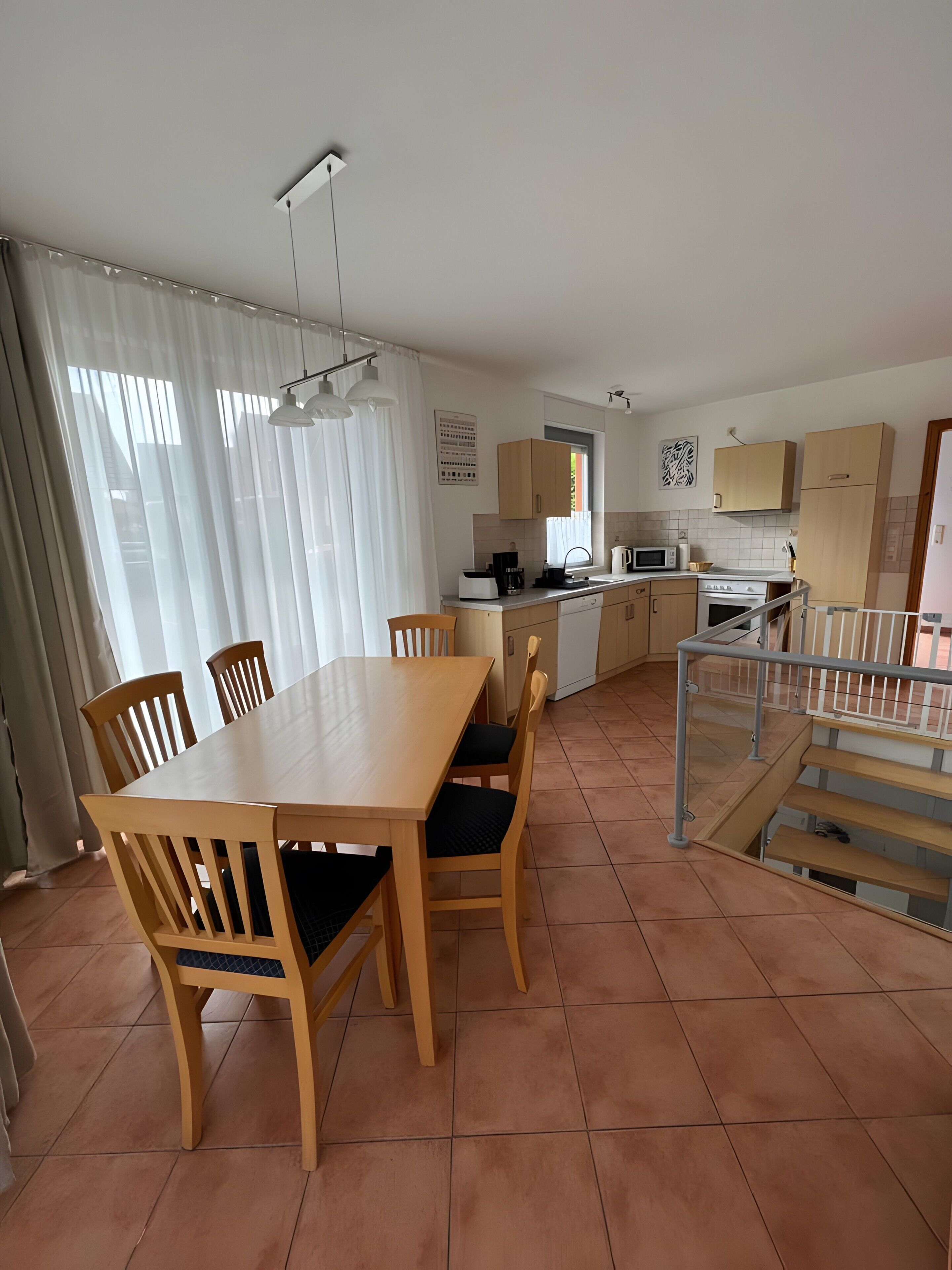 Una cucina con zona pranzo, comprensiva di tavolo e sedie, una scala e una finestra con tende.