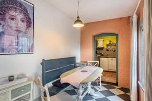 Una habitación con piso a cuadros, una mesa con mantel, un banco, una silla, una lámpara colgante, un cuadro en la pared y una cocina visible a través de un arco.