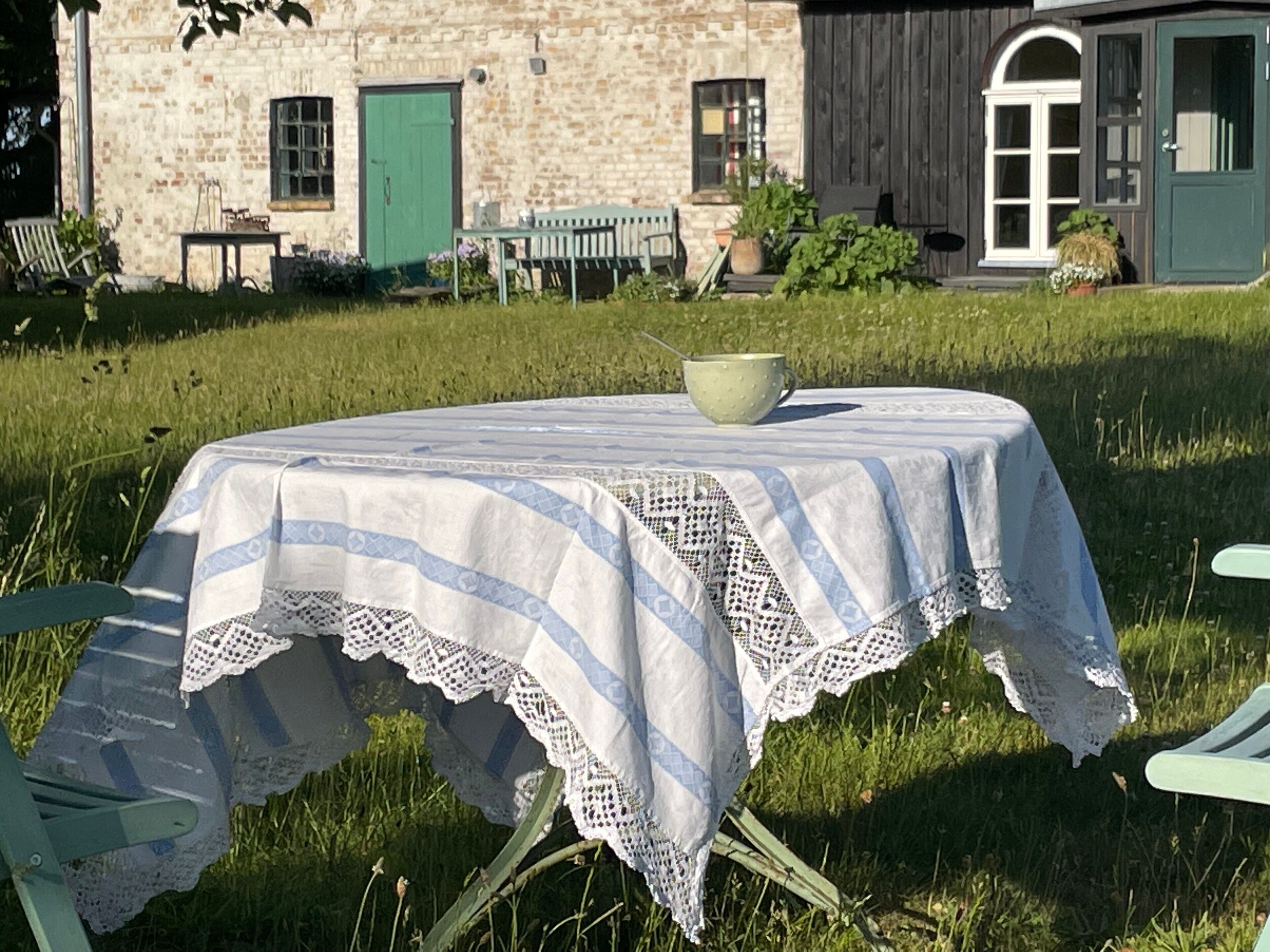 Un bâtiment rustique en pierre, avec une porte et des fenêtres vertes, une table recouverte d’une nappe en dentelle et une chaise verte.