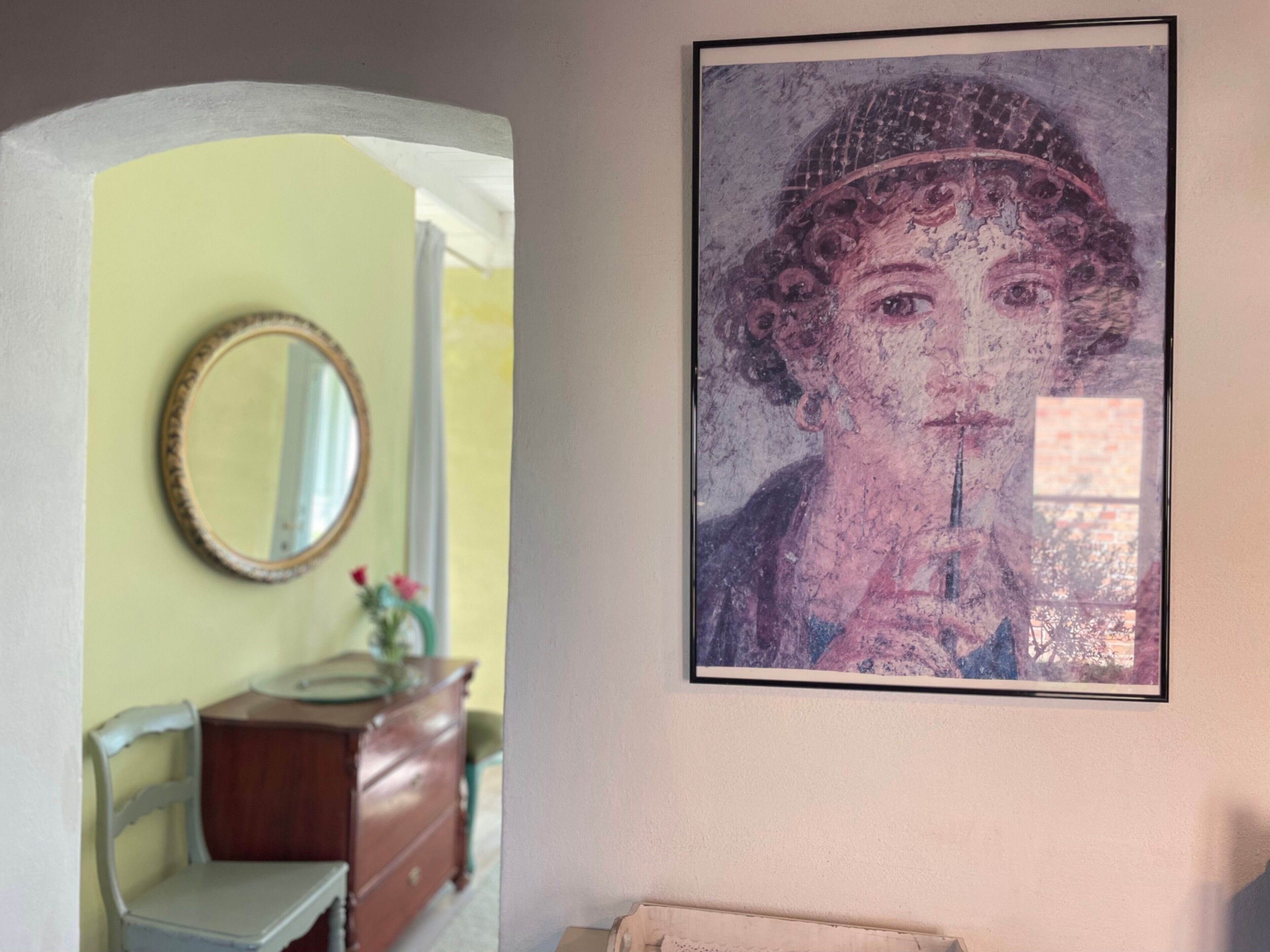 A framed artwork on a wall, a mirror on the wall, and a wooden dresser with a vase of flowers.
