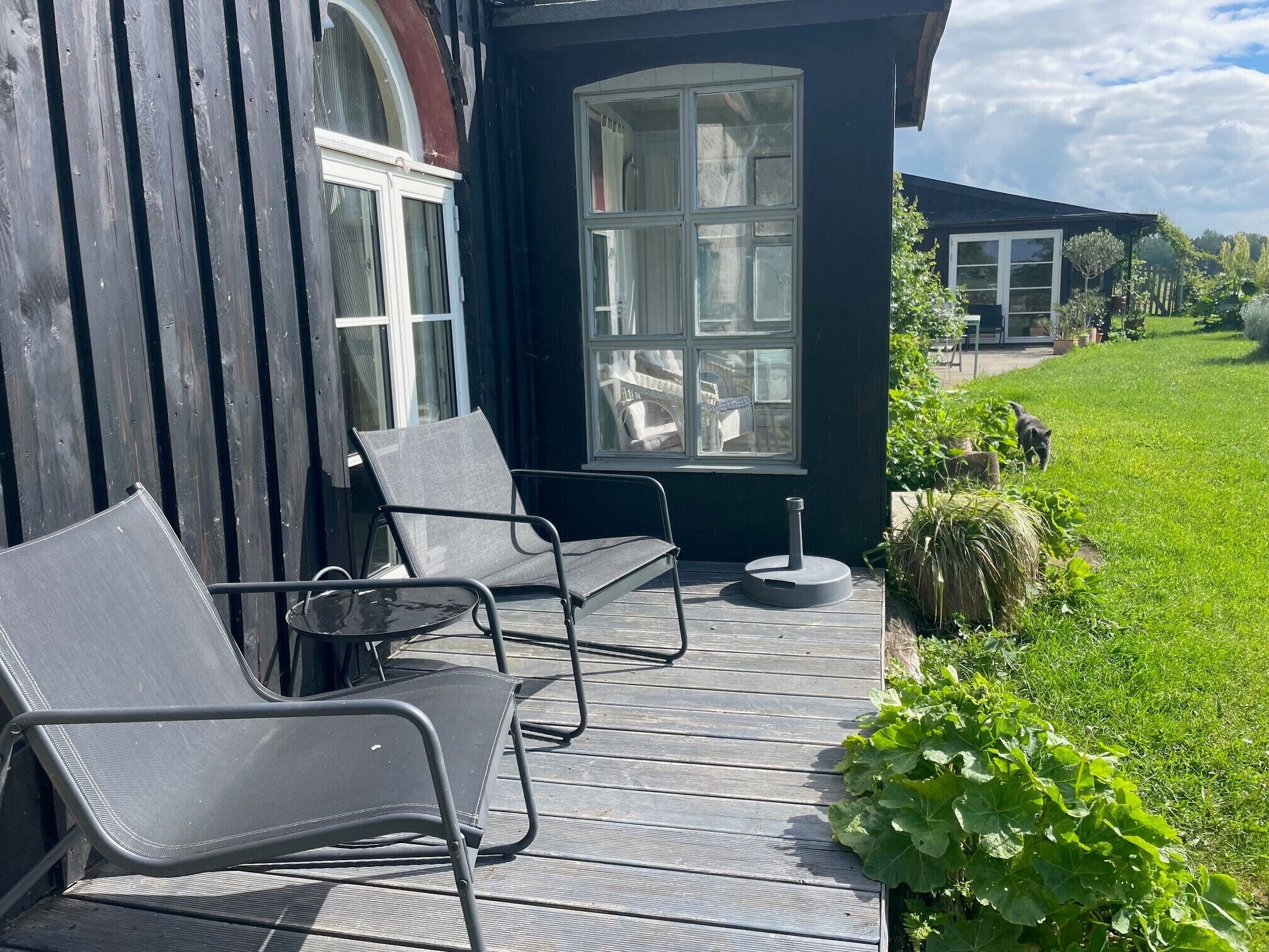 Eine Holzterrasse mit zwei Stühlen und einem kleinen Tisch, ein großes Fenster und ein Rasenstück.
