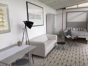 A modern room with a white sofa, a small table, a floor lamp, a bed with striped pillows, and a geometric-patterned rug. - Holiday Home 'Teehaus' (Saal)