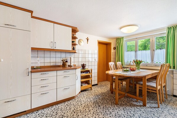 Une cuisine avec des meubles blancs, un plan de travail en bois, un sol carrelé, une table à manger avec des chaises et une fenêtre avec des rideaux.