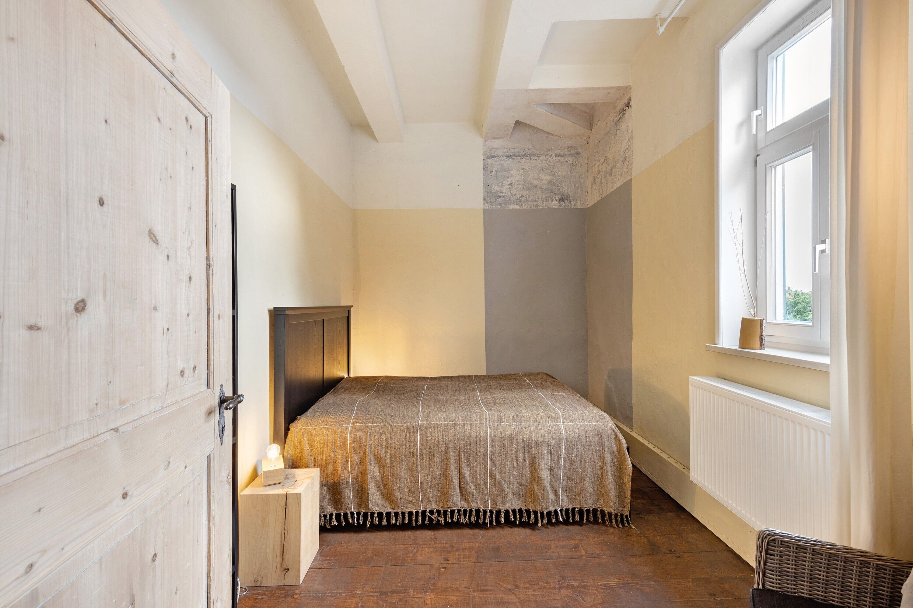 A bedroom with a bed, a window, a radiator, a wooden wardrobe, and a small table with a lamp.