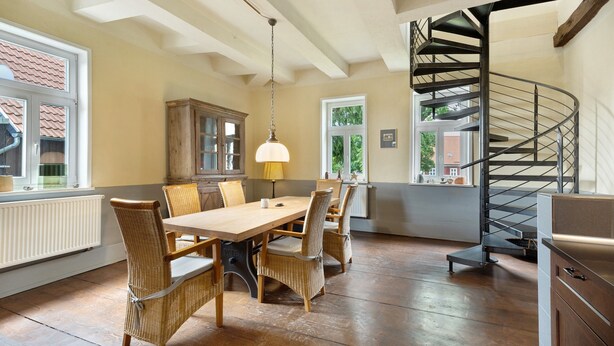 Un espace repas avec une table et des chaises en bois, une lampe suspendue et un escalier en colimaçon.