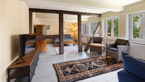 A spacious living area with a large sofa, a coffee table, and a patterned rug. There are two wicker chairs, a wooden bench, and a television on a stand. The room has a high ceiling with exposed beams and large windows. - Apartment 'Altes Pfarrhaus - Durchblick' with Private Garden and Wi-Fi (Hüttenrode)