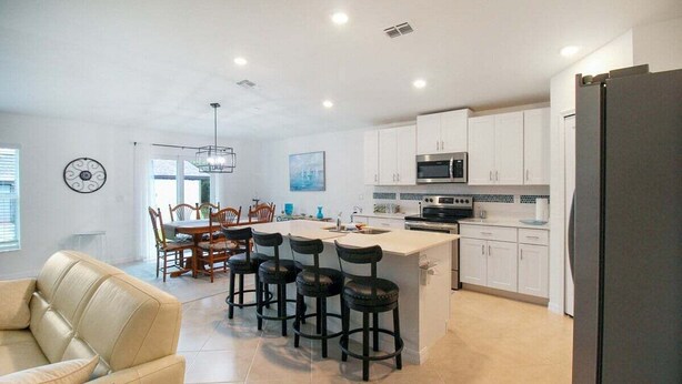 A modern kitchen with white cabinets, a central island with bar stools, a dining area with a table and chairs, and a large refrigerator. - Relaxing coastal home centrally located in Port Charlotte - Fireside Cottage (Port Charlotte)