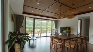 Een ruime eethoek met een grote houten tafel en stoelen, een potplant en uitzicht op een weelderig groen landschap door grote glazen schuifdeuren.