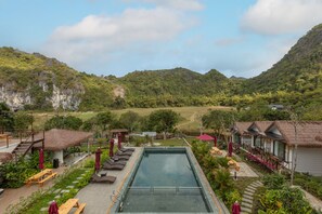 A resort with a swimming pool, lounge chairs, and thatched-roof buildings surrounded by lush greenery and mountains.