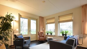 A modern living room with a sofa, armchairs, a coffee table, and a large window with curtains. - Rural apartment with garden (Zuidwolde)