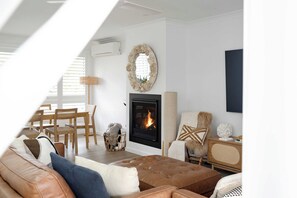 A cozy living room with a fireplace, a dining area with wooden furniture, and a large window providing natural light. - 34° Degrees South Culburra - 4 min walk to beach (Culburra Beach)