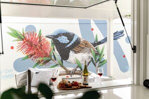 A dining area with a mural of a bird and foliage, a table with a bottle and glasses, and a view of the street.
