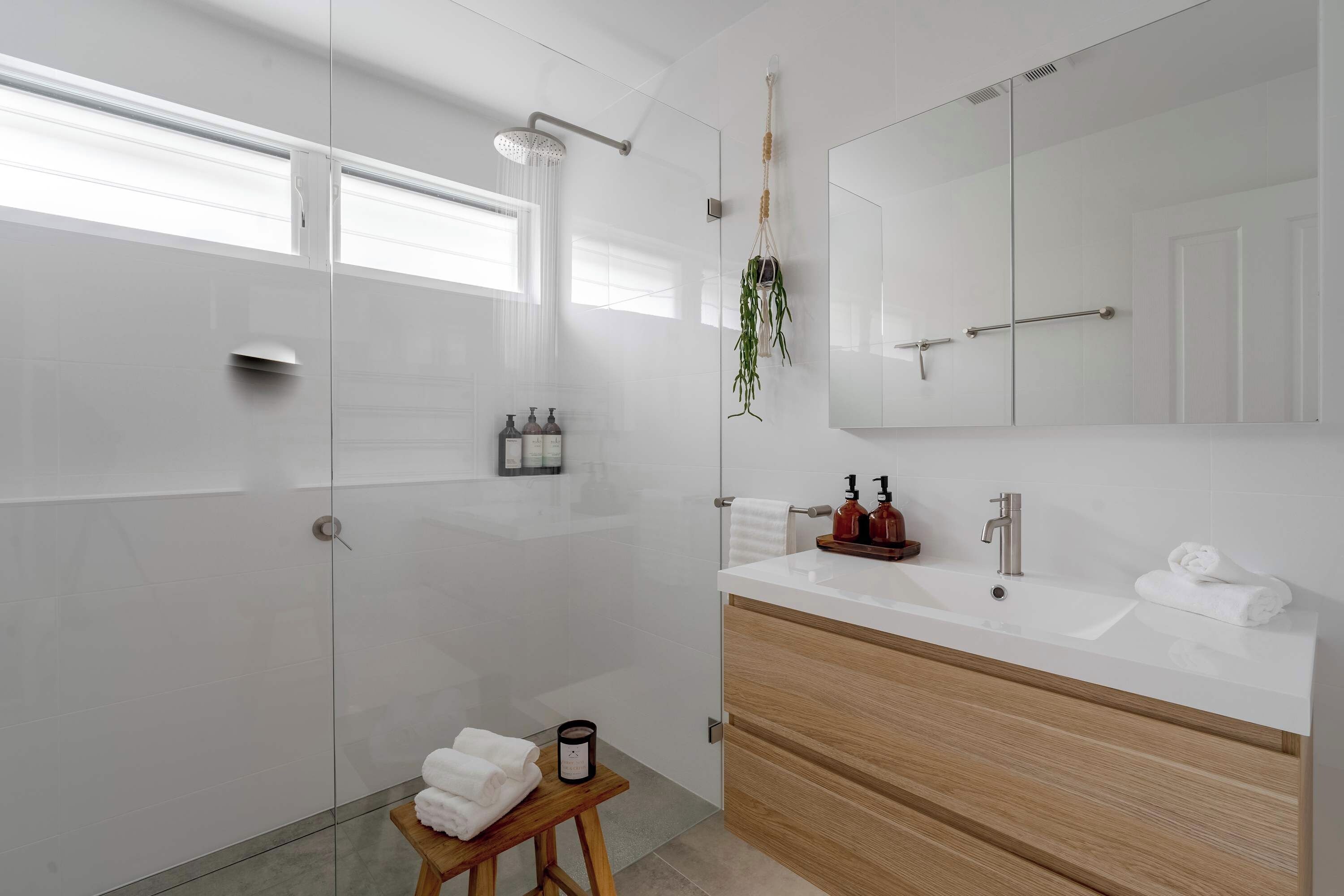 Un baño moderno con cabina de ducha de vidrio, tocador de madera, espejo grande y un taburete pequeño con toallas.