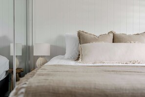 Una cama bien hecha con edredón blanco y beige, una lámpara de noche y una mesita de madera.
