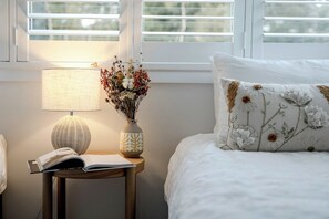 Una cama bien hecha con un cojín floral, una lámpara de noche y un florero con flores secas sobre una mesita de noche.