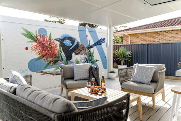 An outdoor patio area with wicker furniture, a mural of a bird and flowers, and a brick building in the background.