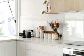 Une cuisine avec des meubles blancs, une machine à café, un vase contenant des fleurs séchées et une étagère avec des livres et des objets décoratifs.