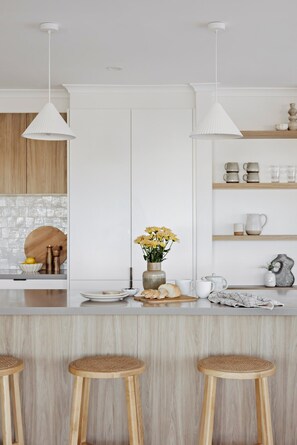 A modern kitchen with white cabinetry, a central island with stools, pendant lighting, and a variety of kitchenware on shelves. - The Cali Jervis Bay (Vincentia)