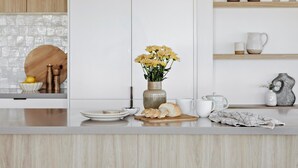 A modern kitchen with white cabinetry, a central island with stools, pendant lighting, and a variety of kitchenware on shelves. - The Cali Jervis Bay (Vincentia)