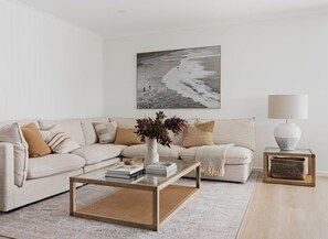 A modern living room with a beige sofa, a glass coffee table, a framed beach photograph, a vase with flowers, and a lamp on a side table.
