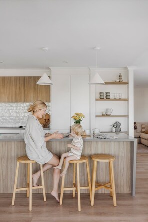 A modern kitchen with wooden cabinets, a central island with stools, pendant lighting, and a backsplash. - The Cali Jervis Bay (Vincentia)