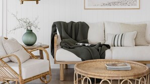 A living room with a white sofa, a wicker coffee table, a wicker armchair, a hanging light fixture, and a framed picture on the wall. - The Cali Jervis Bay (Vincentia)
