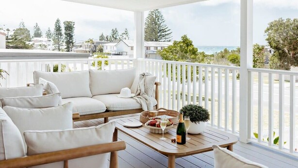 A covered outdoor seating area with white furniture, a wooden table, and a view of the ocean. - The Cali Jervis Bay (Vincentia)