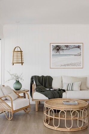 A living room with a white wall, a wicker coffee table, a wicker armchair, a sofa with a green throw blanket, a hanging light fixture, and a framed picture of a beach scene. - The Cali Jervis Bay (Vincentia)