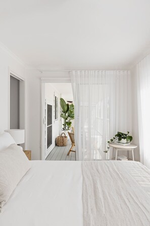 A minimalist bedroom with a large bed, a small table with a plant, and a view through a doorway to an outdoor area with plants and a wooden floor.