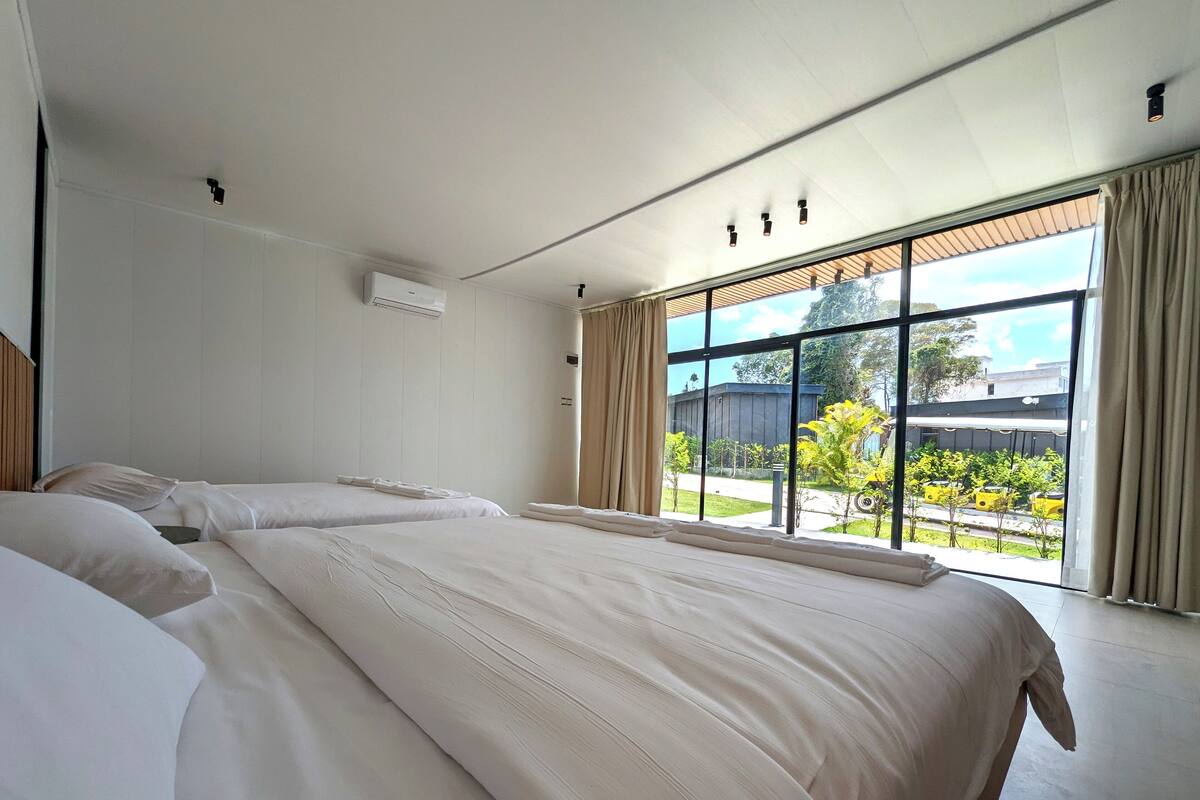 A modern hotel room with two beds, a large window with curtains, and a view of the outside.