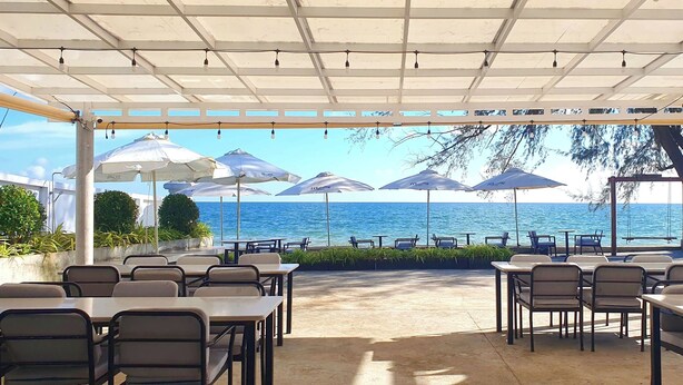 A covered outdoor seating area with tables and chairs, umbrellas, and a view of the sea.