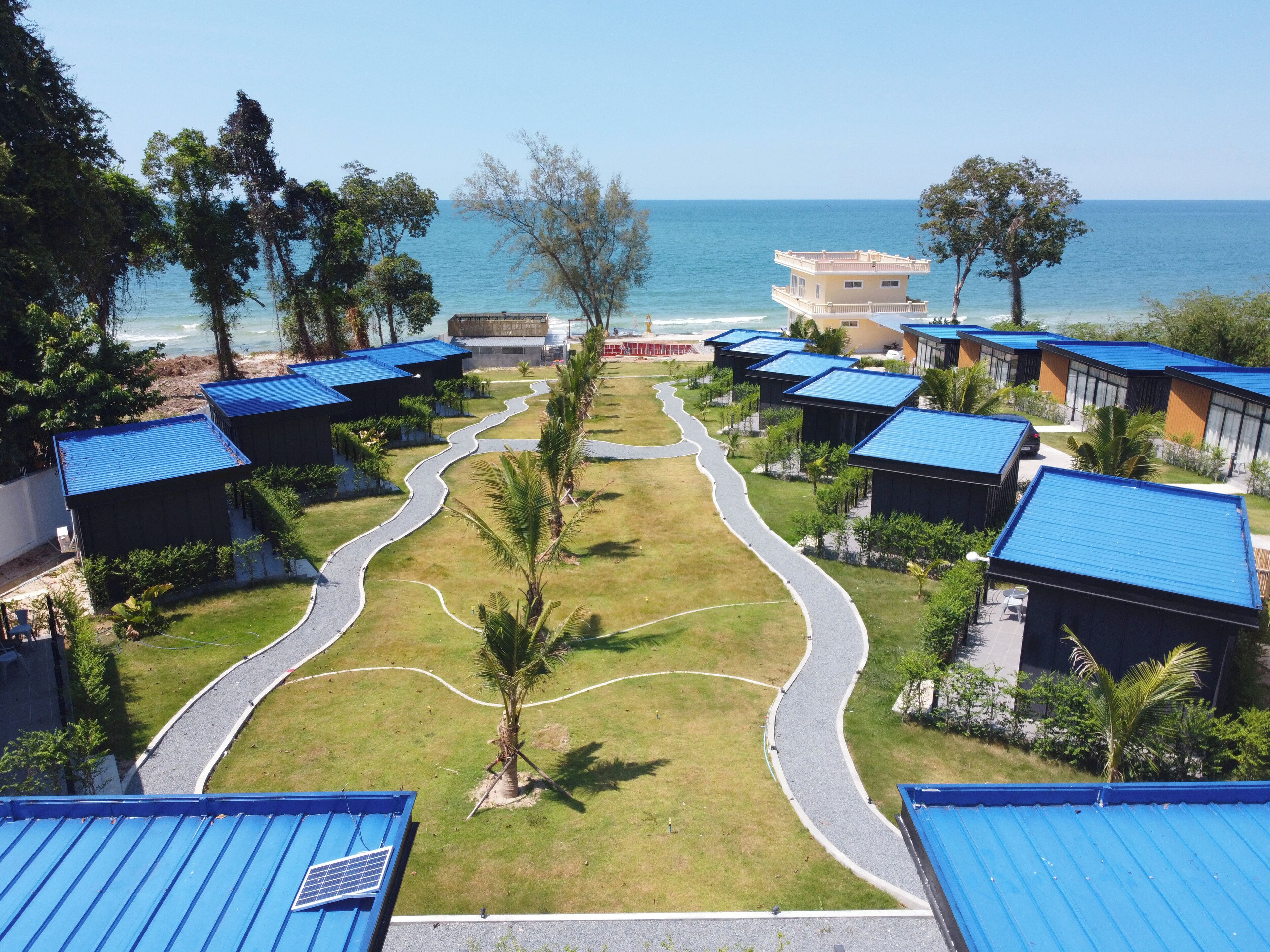 A resort with multiple blue-roofed buildings arranged in a curved layout, a central pathway, and a view of the sea.
