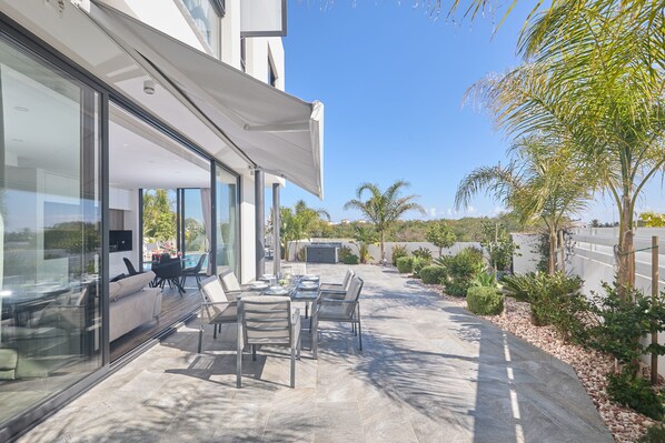 A modern outdoor patio with a glass enclosure, a dining table with chairs, and a pool in the background.