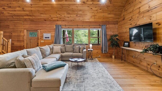 A cabin interior with a sectional sofa, a TV, a window with curtains, and a wooden ceiling.